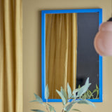 Atle rectangular wall mirror, 40x60cm, with a narrow blue MDF frame, shown on a beige wall reflecting gold curtains, with a hand adjusting it and a plant in foreground.