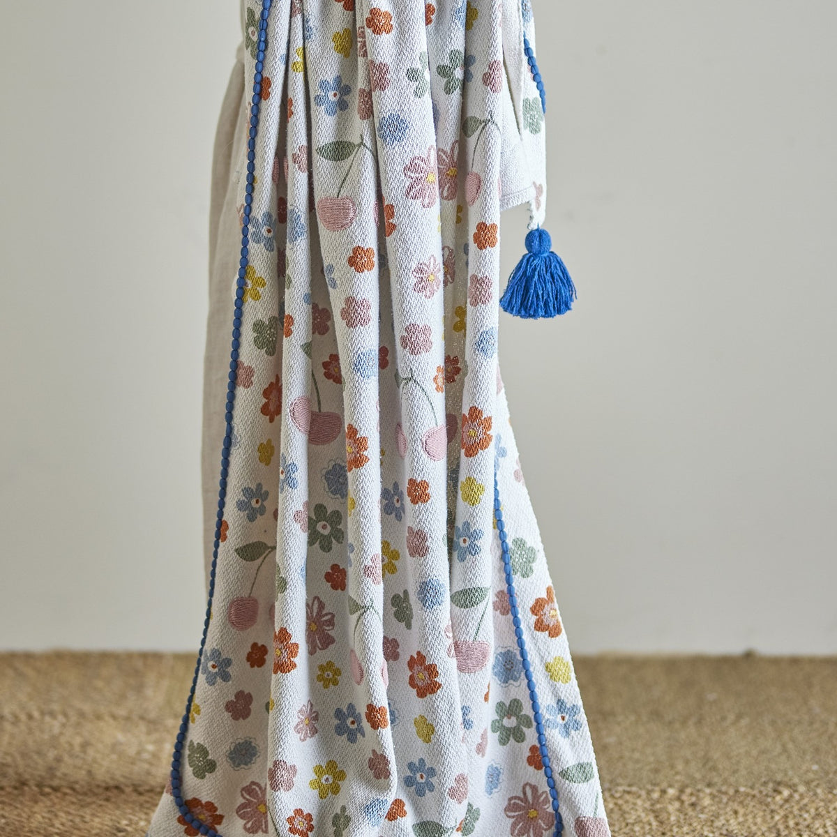 Cherry Floral Cotton Throw in multicoloured woven cotton with a floral and cherry print, finished with a blue pom-pom trim and tassel, draped against a plain background.