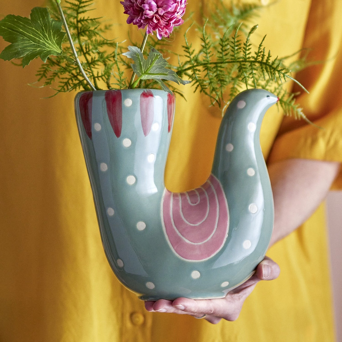 Trudy Bird Stoneware Vase in green, held in hand, showing its curved neck, polka-dot body, pink wing detail, and raised tail opening holding flowers and foliage.