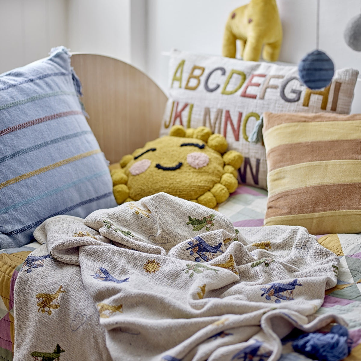 Plane Cotton Throw in beige cotton blend, patterned with biplanes, clouds and suns, draped on a nursery bed beside striped and alphabet cushions.