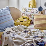Plane Cotton Throw in beige cotton blend, patterned with biplanes, clouds and suns, draped on a nursery bed beside striped and alphabet cushions.