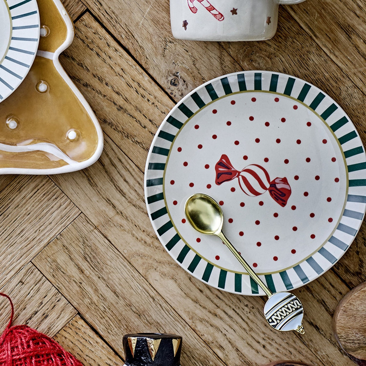 Yule Striped Stoneware Christmas Plate in multicoloured ceramic, featuring a green and white striped rim, red polka dots, and a wrapped sweet design, shown with a gold spoon on a wooden table.