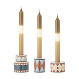 Bitja Geometric Christmas Candle Holders, set of 3 round stoneware bases in multicoloured zigzag, triangle and diamond patterns, holding lit taper candles on a plain white background.