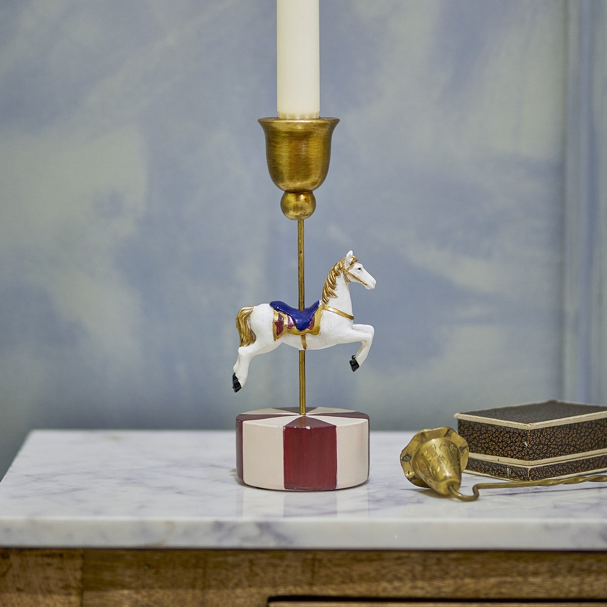 Merry-go-round resin Christmas candle holder with a hand-painted white carousel horse on a brass rod, round red and pink striped base, shown on a marble surface with a lit candle.