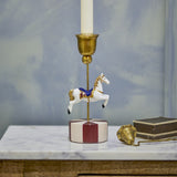 Merry-go-round resin Christmas candle holder with a hand-painted white carousel horse on a brass rod, round red and pink striped base, shown on a marble surface with a lit candle.