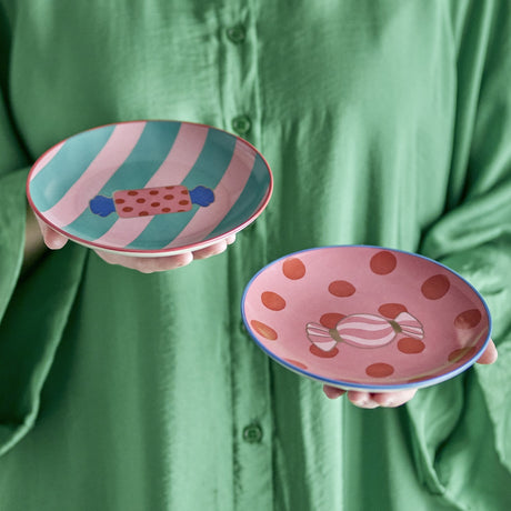 Set of 2 Caramella stoneware Christmas plates held in hands, featuring pink, teal, and red stripe and dot sweet motifs with contrasting rim bands.