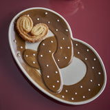 Stoneware serving platter shaped like a pretzel, glossy brown with white dot detailing, holding two sugared pastry rolls, set on a dark red background.