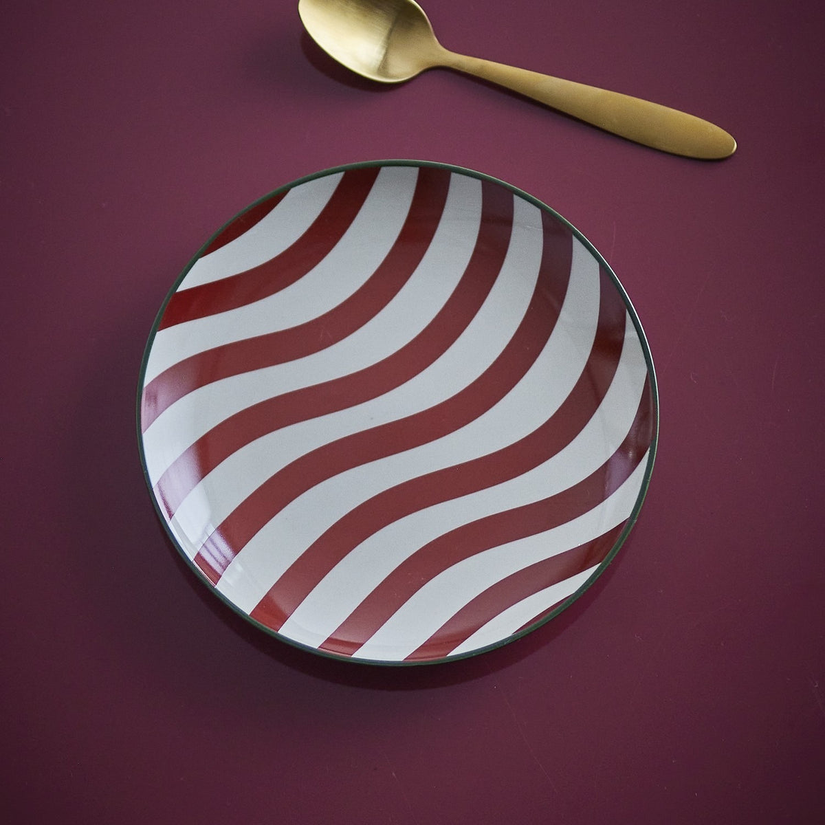 Jolly Striped Stoneware Christmas Plate in red and white wavy stripes with a thin rim line, shown beside a gold spoon on a dark burgundy surface.
