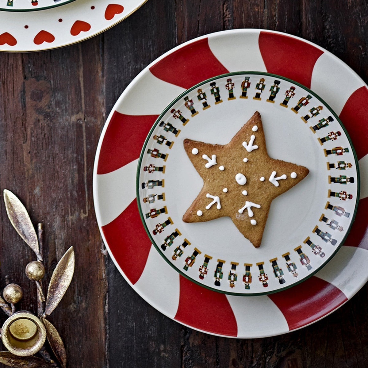 Jolly Stoneware Christmas Side Plate in cream, featuring a nutcracker soldier rim, shown stacked on a red and white striped plate with a star-shaped iced biscuit on a dark wood table.