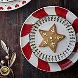 Jolly Stoneware Christmas Side Plate in cream, featuring a nutcracker soldier rim, shown stacked on a red and white striped plate with a star-shaped iced biscuit on a dark wood table.