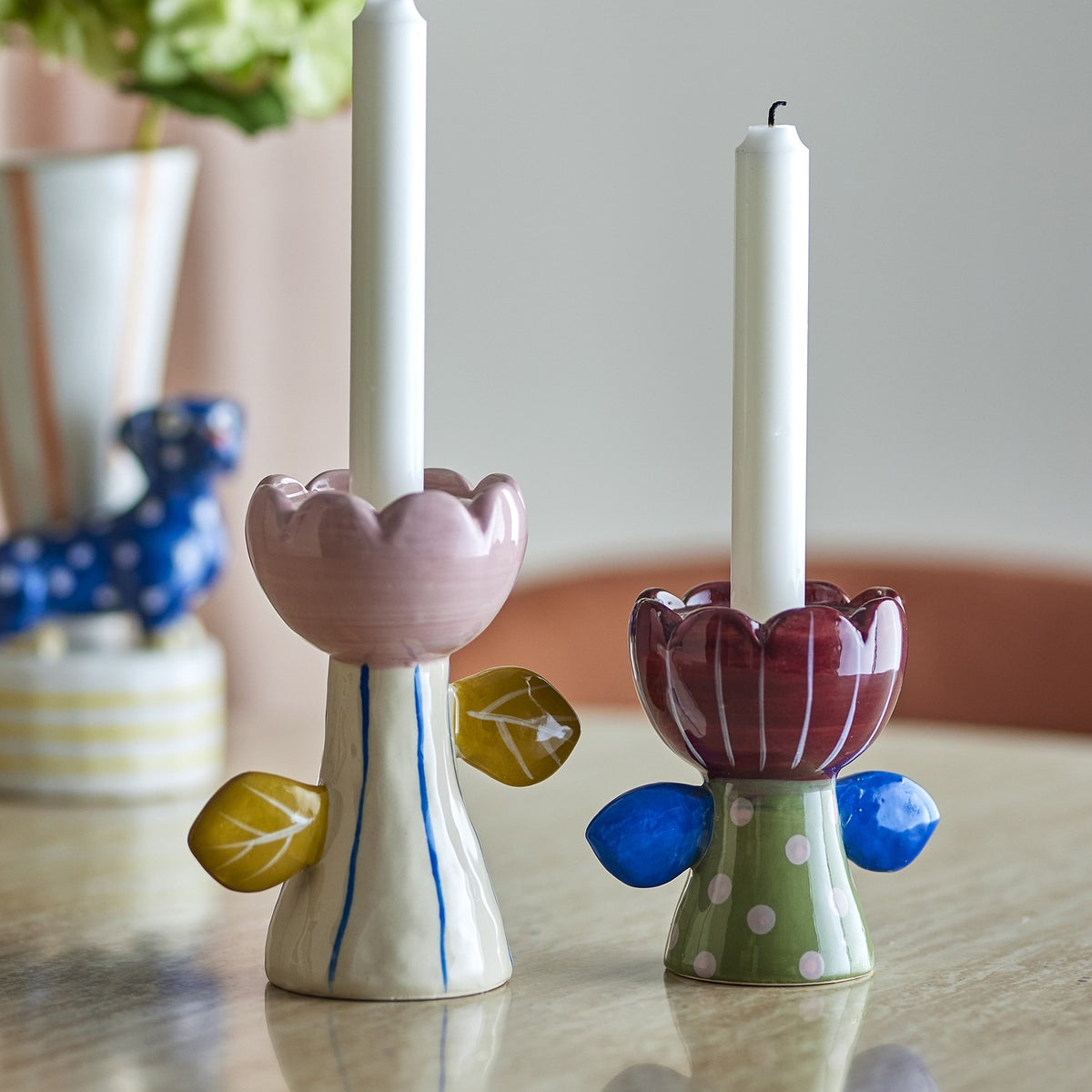 Set of 2 handpainted ceramic Florita flower candle holders with scalloped petal cups and leaf details, in pink with yellow leaves and burgundy with blue leaves, holding white candles.