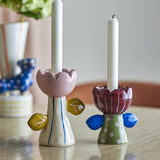 Set of 2 handpainted ceramic Florita flower candle holders with scalloped petal cups and leaf details, in pink with yellow leaves and burgundy with blue leaves, holding white candles.