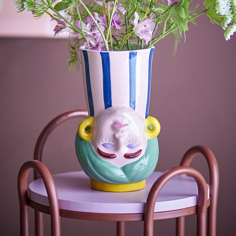 Miss Jody Stoneware Vase 22.5cm in Multi, a ceramic face-shaped vase with closed eyes, yellow earrings, teal hair base, and a blue-striped neck, holding pink florals on a lilac stool.