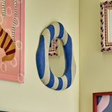 Travis Striped Resin Wall Mirror in blue and white, an irregular rounded shape with a cushioned striped frame, hung on a pale yellow wall beside framed artwork.