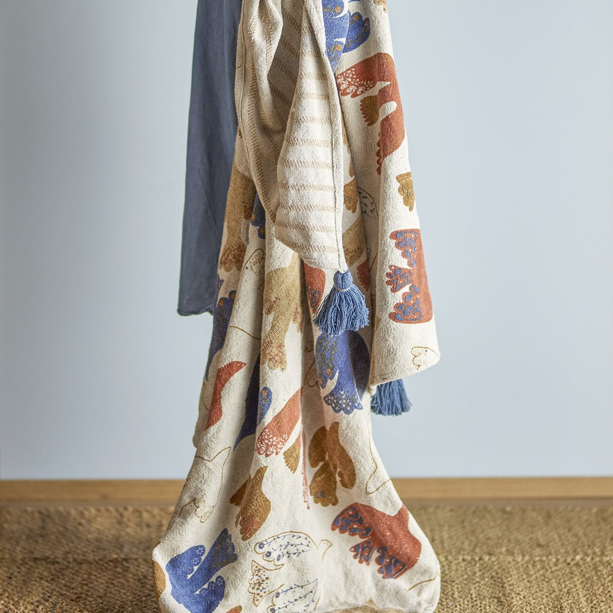 Anniversary Cotton Throw in Multi, draped showing a textured weave with bird and wing print in rust, blue and ochre tones, finished with a blue corner tassel, against a light grey background.