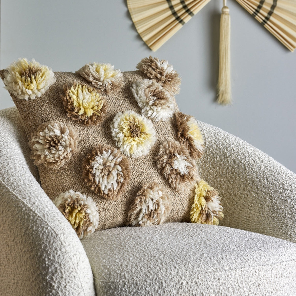 Somma Wool Cushion in Natural with tufted shaggy rosettes in beige, cream, and yellow, shown on a bouclé armchair against a grey wall.