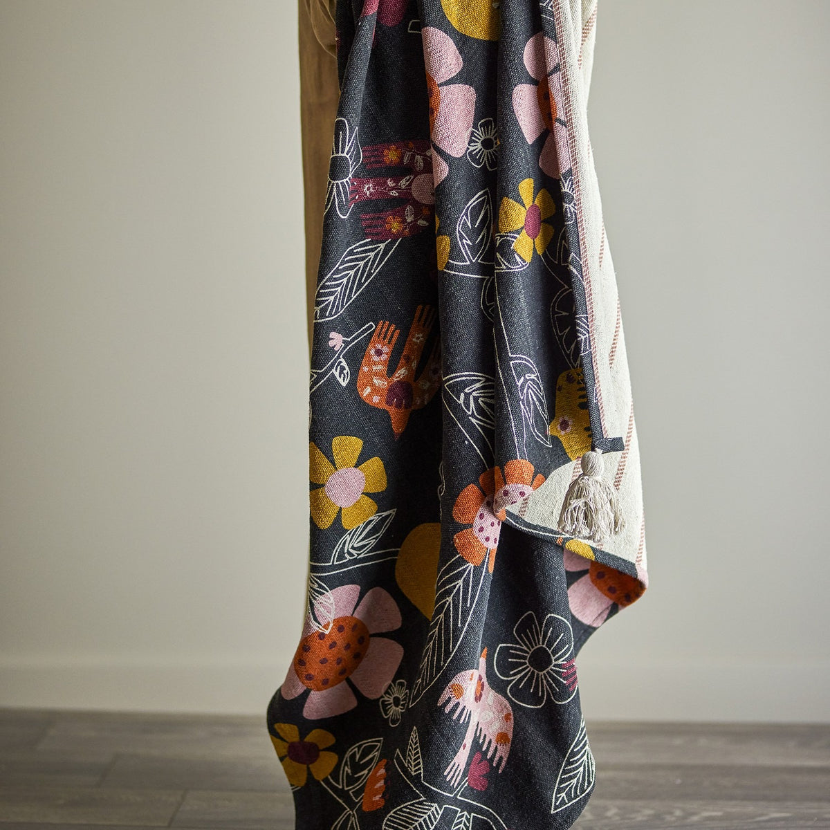 Vivielle Striped Throw draped over a chair, showing its multicoloured floral and bird print on a dark ground, with the striped reverse side visible at the corner fold.