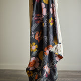 Vivielle Striped Throw draped over a chair, showing its multicoloured floral and bird print on a dark ground, with the striped reverse side visible at the corner fold.