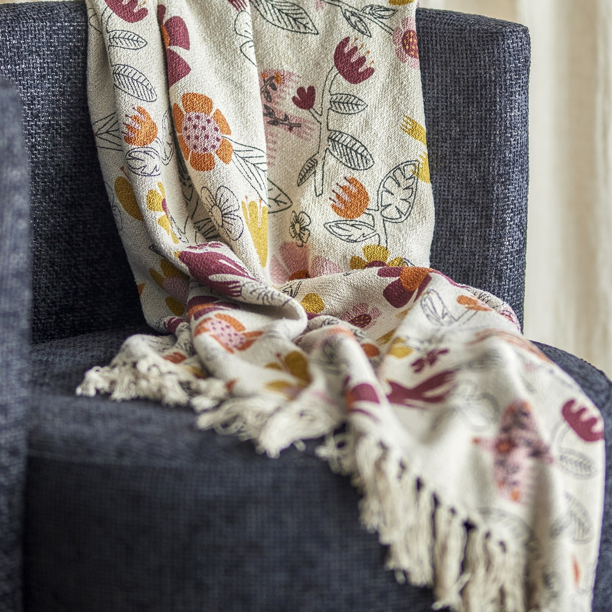 Vivielle Floral Throw in multicoloured cotton-blend fabric with a printed pattern of flowers, leaves and birds and fringed edges, draped over a dark grey armchair.
