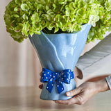 Belivia Bouquet Stoneware Vase 28.5cm in blue, with a wavy rim, draped folds, and a glossy polka-dot bow at the base, held in hands beside green hydrangea blooms.