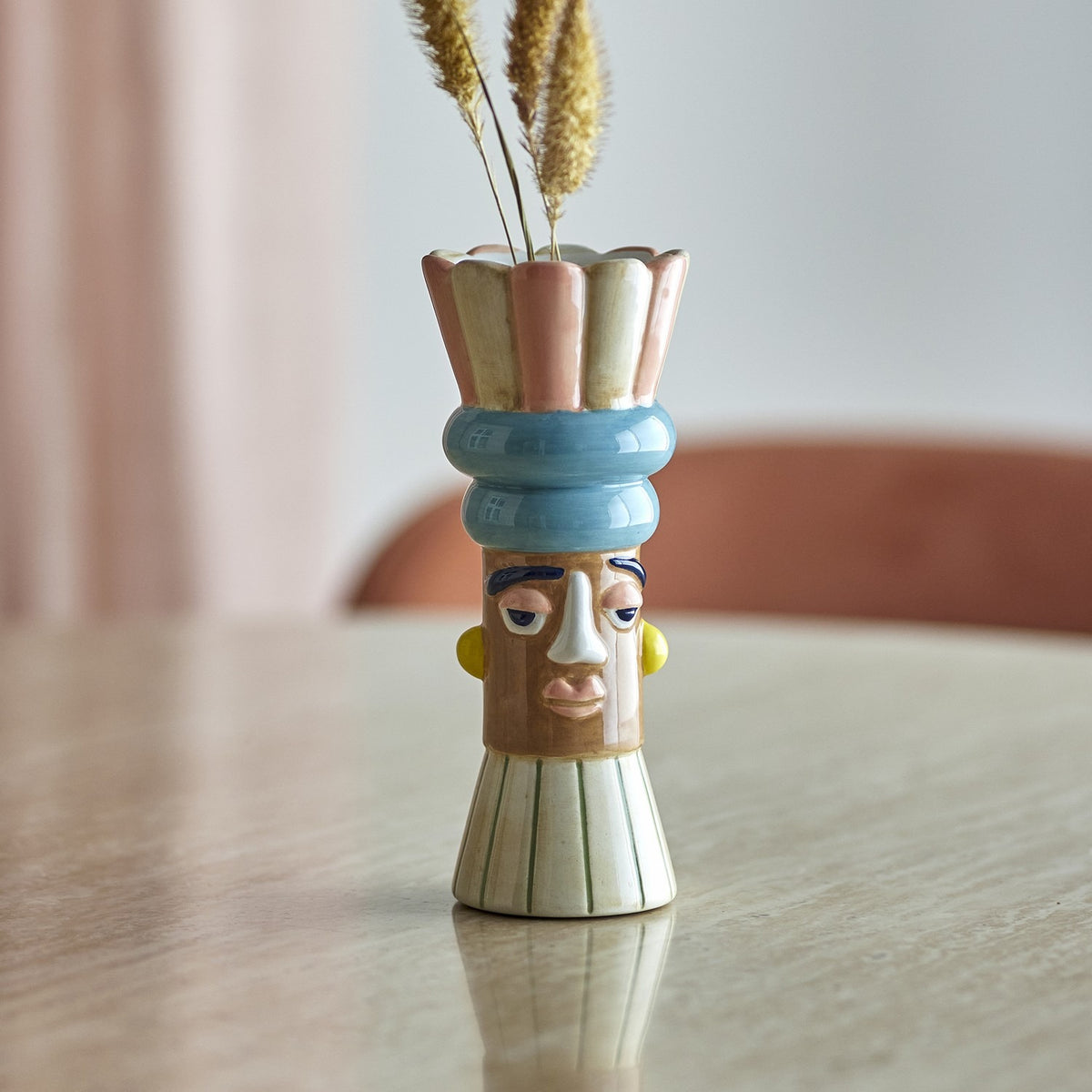 Minio Ceramic Vase 17cm, a multicoloured face-design bud vase with scalloped petal rim and blue stacked headdress bands, holding dried grass stems on a table.