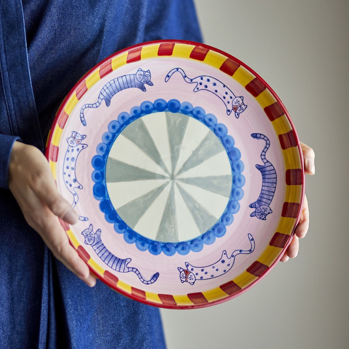 Missy Striped Stoneware Tray held in hand, round ceramic serving dish with blue cat motifs, scalloped blue centre ring, and red and yellow striped rim.