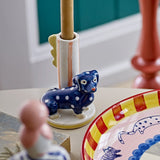 Dachsy Dog Stoneware Candle Holder in multicoloured ceramic, featuring a polka-dot dachshund beside a striped candle tube with scalloped fin, shown on a table with decorative plate and figurine.