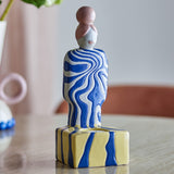 Priya Stoneware Tealight Holder in Multi, a ceramic figure with a bun hairstyle and blue-and-white swirl-patterned body, standing on a striped yellow and blue base with a round tealight recess.