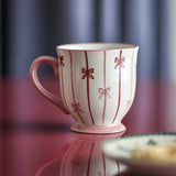 Hope Striped Stoneware Christmas Mug in white with pink stripes and bow details, footed base and curved handle, shown on a glossy pink surface.
