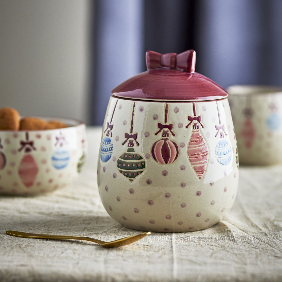 Ornamentia Dotted Christmas Storage Jar with Lid 890ml in cream stoneware, featuring hand-painted hanging baubles and dots, with a burgundy glazed lid and bow-shaped knob.