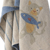 Mille Cotton Throw in beige woven cotton, close-up of a printed cat holding a fish design with a striped blue border and reverse side stripe detail.