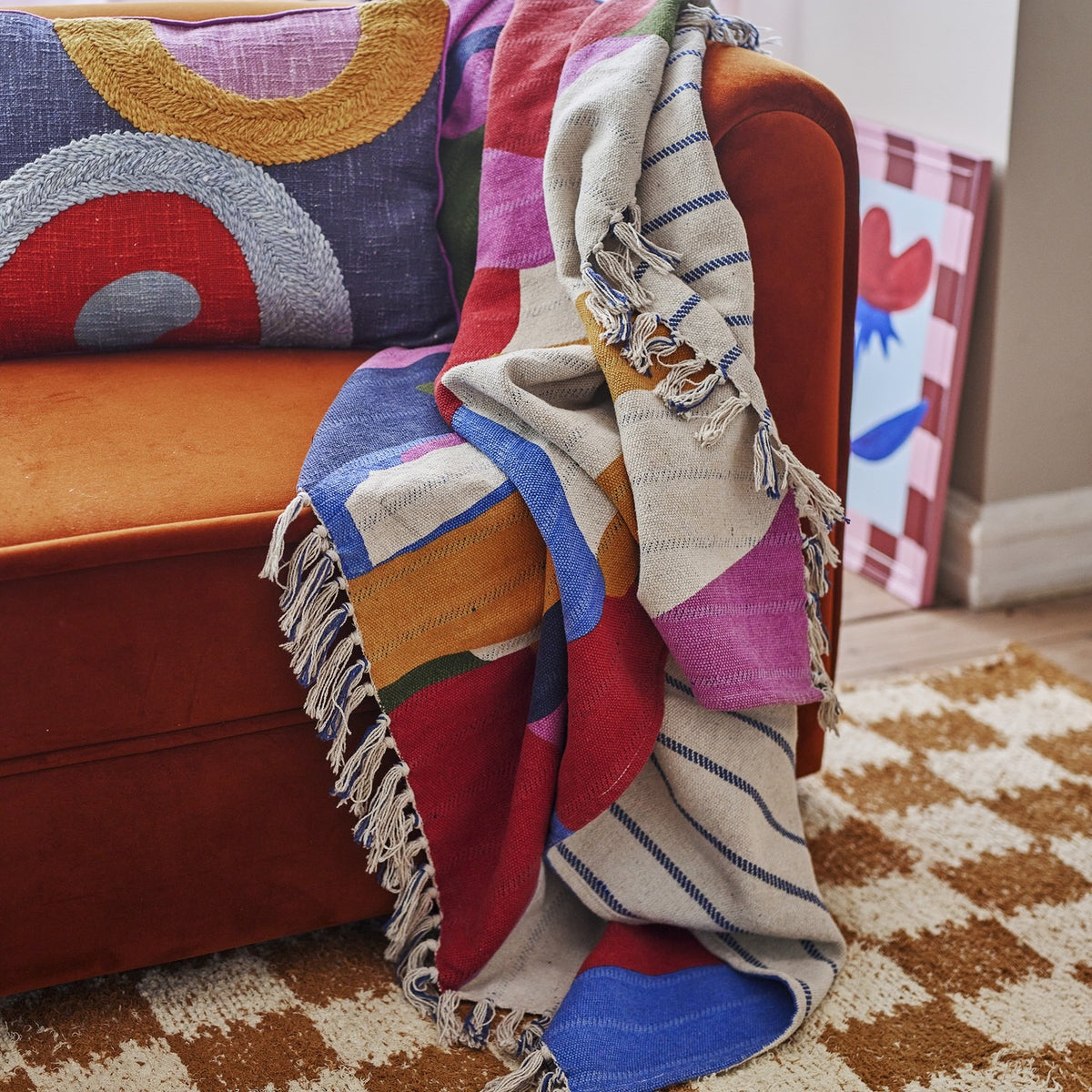 Artista Cotton Throw in multicoloured abstract print with knotted fringe, draped over an orange velvet sofa in a living room setting.