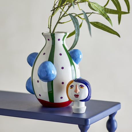 Conneli Ceramic Vase 33cm in white with green stripes, purple dots, red base and blue knobs, paired with a matching figural head stopper, styled with foliage on a dark blue table.