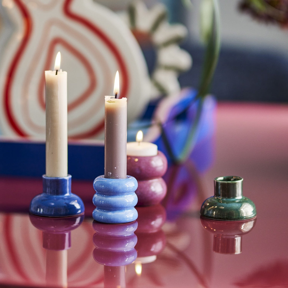 Tana Speckled Tealight & Candle Holders Set of 2 in ribbed ceramic with reactive glaze, shown in blue and pink tones holding lit candles, with a green holder alongside on a glossy tabletop.