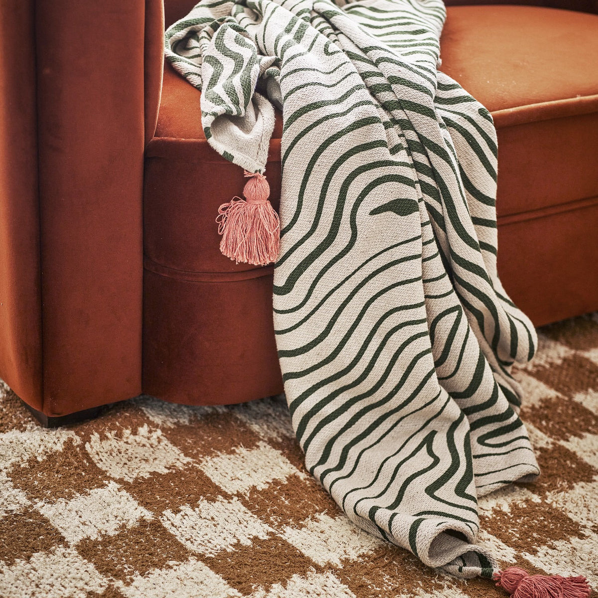 Woodsby Striped Cotton Throw in white and green, draped over a rust velvet sofa with a pink tassel corner visible, set on a patterned rug.