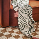 Woodsby Striped Cotton Throw in white and green, draped over a rust velvet sofa with a pink tassel corner visible, set on a patterned rug.