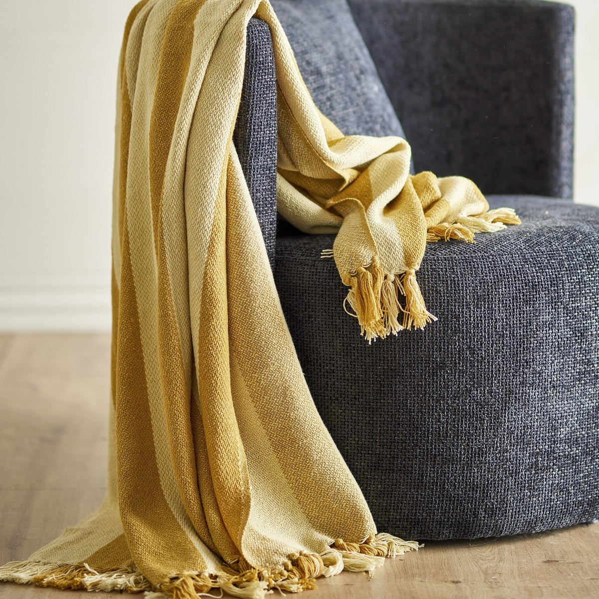 Frello Striped Cotton Throw in yellow with alternating deep and pale stripes and fringed edges, draped over a dark grey upholstered armchair.
