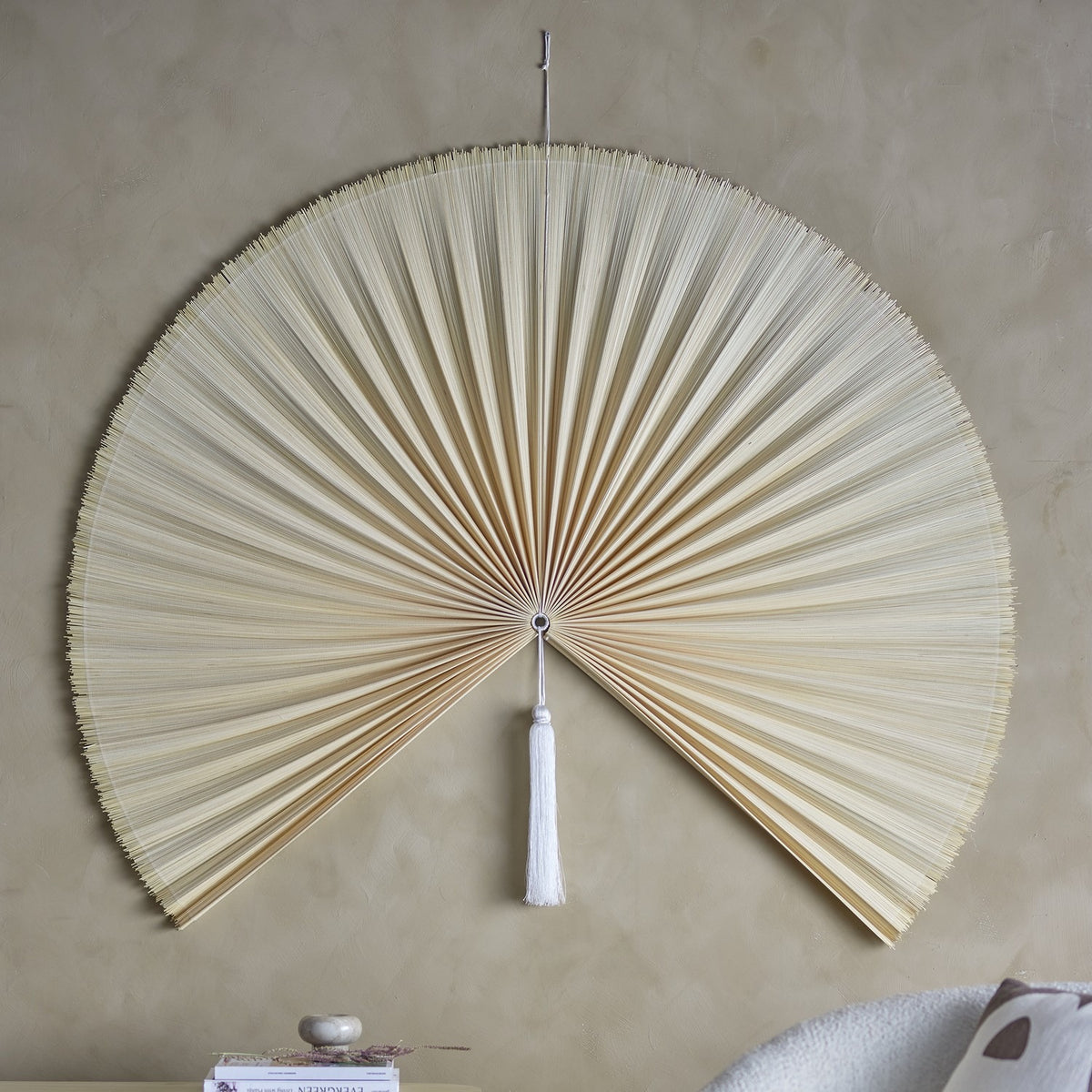 Jaime Bamboo Wall Decor 110cm in Natural, a large pleated bamboo fan-shaped wall art piece with a white tassel at centre, mounted on a neutral wall above a shelf with books.