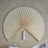 Jaime Bamboo Wall Decor 110cm in Natural, a large pleated bamboo fan-shaped wall art piece with a white tassel at centre, mounted on a neutral wall above a shelf with books.