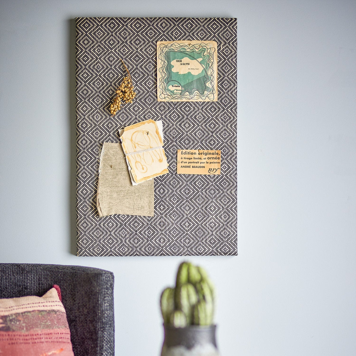 Rivalta Pin Board in black with woven diamond pattern, shown mounted on a wall with pinned postcards, dried flowers, and fabric swatch above a sofa.
