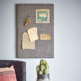 Rivalta Pin Board in black with woven diamond pattern, shown mounted on a wall with pinned postcards, dried flowers, and fabric swatch above a sofa.