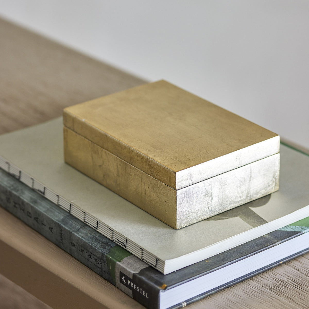 Nenon rectangular storage box with lid in gold MDF with a high gloss finish, shown atop stacked books on a wooden shelf.