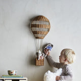 Natural rattan wall basket shaped like a hot air balloon with iron frame, displayed in a nursery as a child reaches for plush elephants inside.