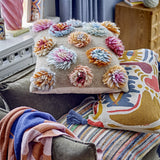 Somma Wool Cushion in Multi with tufted flower-like pompoms in pink, blue and ochre, shown on a sofa alongside a patterned cushion and striped throw.