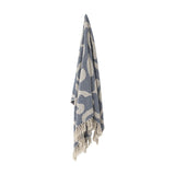 Salobre Cotton Throw in blue, woven with large abstract shapes in a lighter tone, finished with knotted fringe and hung against a plain white background.