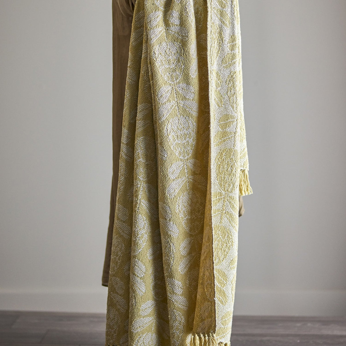 Pratello Throw in yellow woven fabric with a leafy floral pattern and fringed edge, draped over furniture against a plain neutral background.