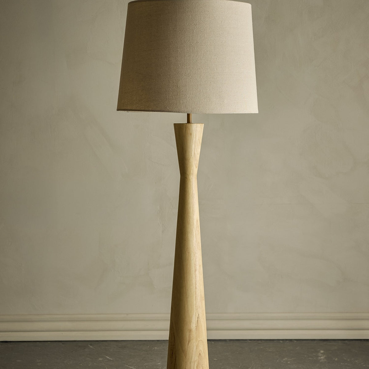 Leonor Wooden Floor Lamp with a natural rubberwood column, narrow waist and tapered linen drum shade, shown against a plain neutral background.