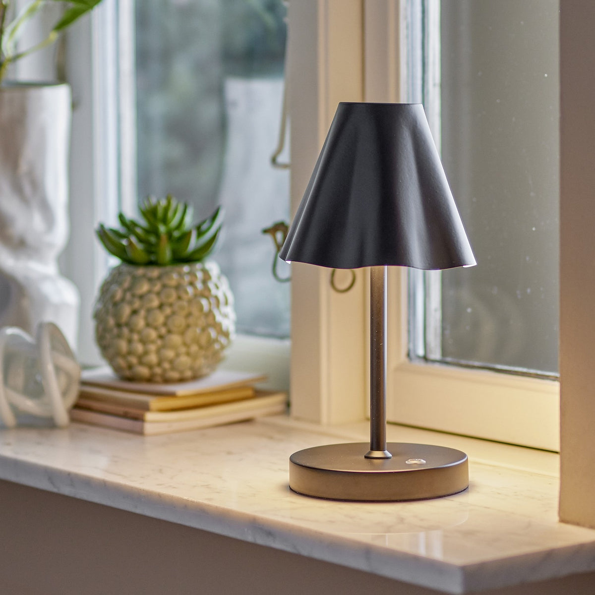 Lianna Wavy Iron Rechargeable Table Lamp in black, with a cone-shaped shade and wavy lower edge on a slender stem, shown lit on a marble windowsill beside books and a succulent.