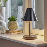Lianna Wavy Iron Rechargeable Table Lamp in black, with a cone-shaped shade and wavy lower edge on a slender stem, shown lit on a marble windowsill beside books and a succulent.
