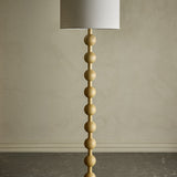 Giacoma Wooden Floor Lamp in natural wood, featuring a stacked column of rounded wooden spheres and a white linen drum shade, set against a plain neutral wall.